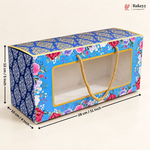 A rectangular bakery gift box with a floral print, blue patterned side panel, clear front window, and gold rope handles. Measurement arrows and labels indicate dimensions of 28 cm length, 10 cm width, and 13 cm height on a light beige background.