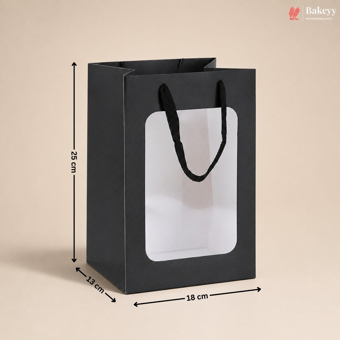 A black paper gift bag with black handles and a large clear front window, shown with dimension arrows and labels indicating 25 cm height, 13 cm width, and 18 cm depth on a beige background.