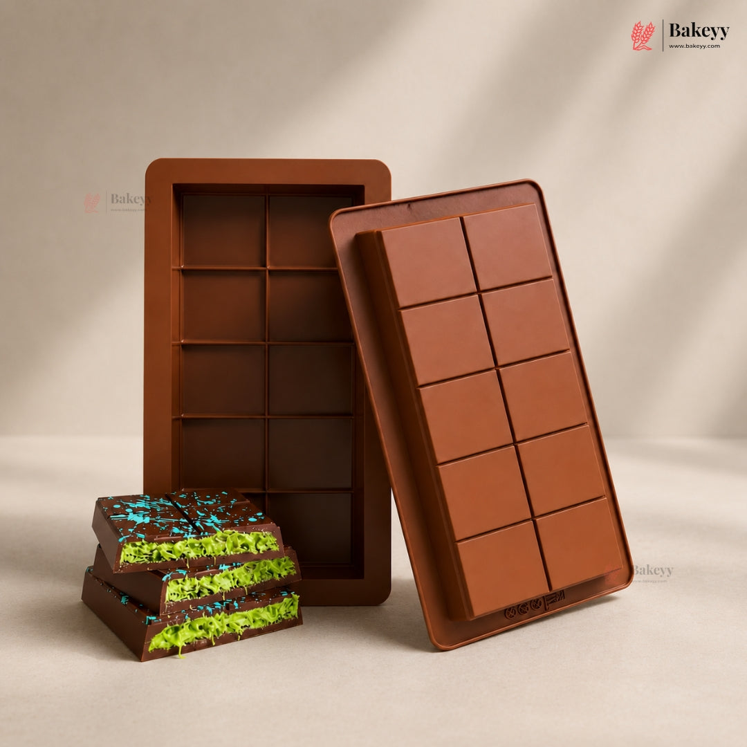 Two brown silicone mold trays and a stack of chocolate bars with bright green and blue sprinkles, displayed on a warm beige background. The Bakeyy brand logo is visible in the top right corner.