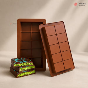 Two brown silicone mold trays and a stack of chocolate bars with bright green and blue sprinkles, displayed on a warm beige background. The Bakeyy brand logo is visible in the top right corner.