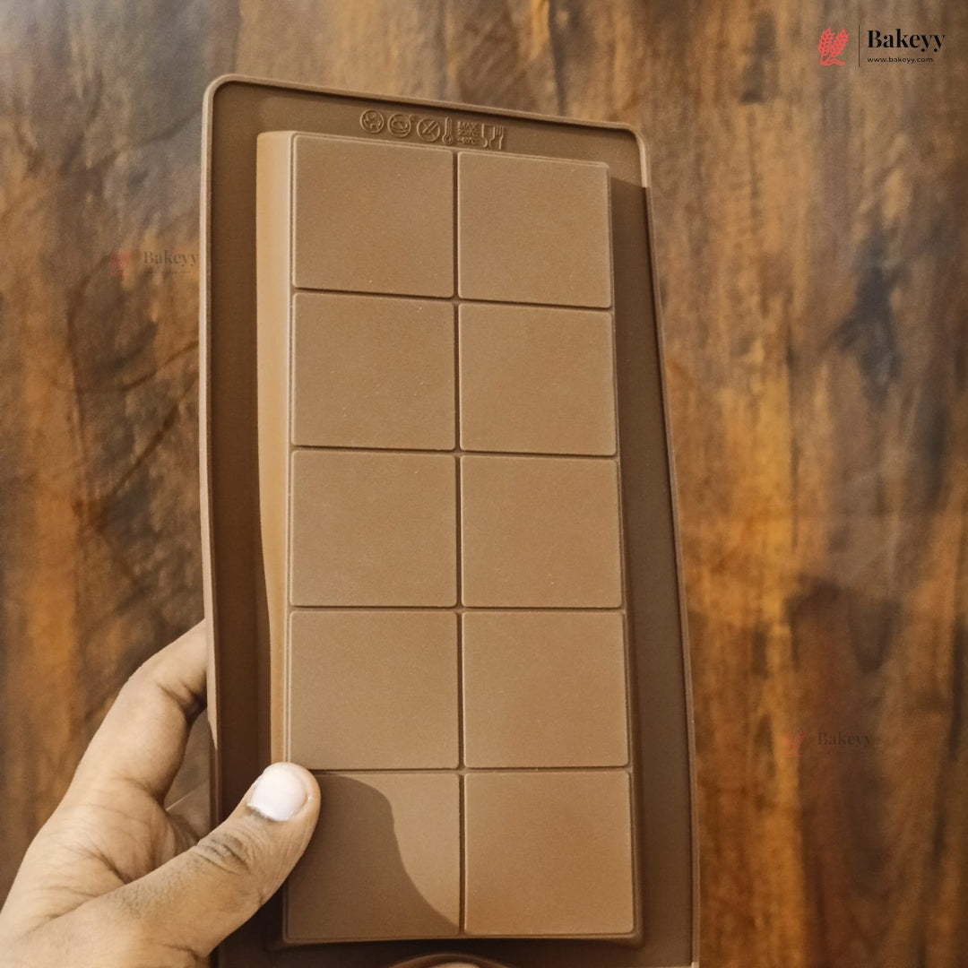 A hand holding a brown silicone mold designed to make eight square chocolate bars, set against a dark wooden background.