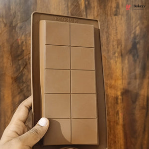 A hand holding a brown silicone mold designed to make eight square chocolate bars, set against a dark wooden background.