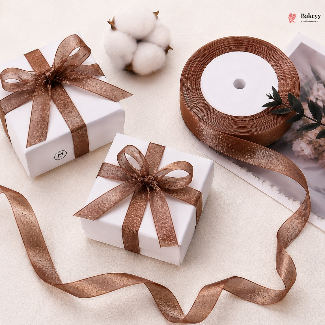 Two white gift boxes with brown ribbons on a light surface, with a roll of brown ribbon and cotton bolls in the background.