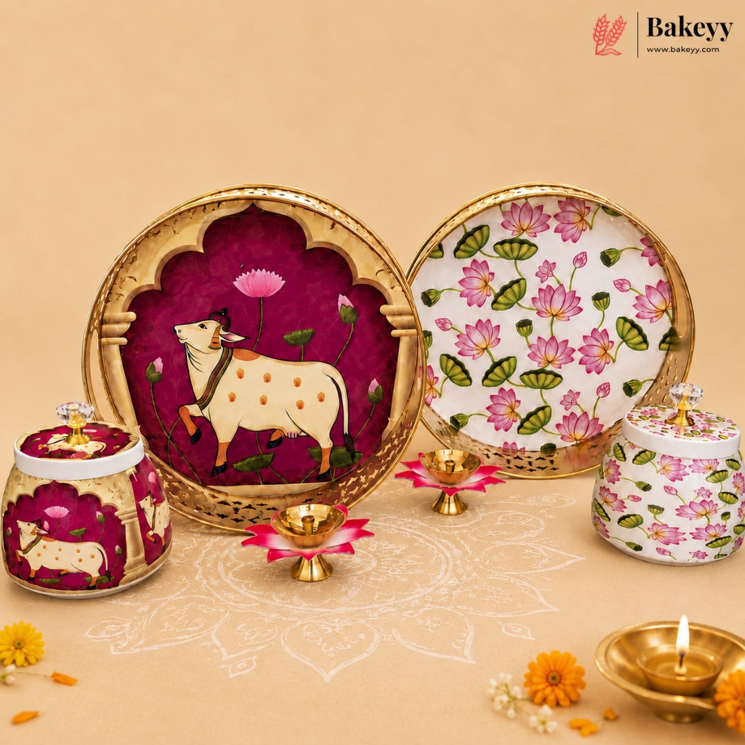 A styled product display of a traditional Indian bakery set by Bakeey, featuring a large round decorative serving platter with a painted cow and lotus motif, a matching patterned plate, and two lidded jars with floral and cow designs, arranged on a beige surface with lit oil lamps.