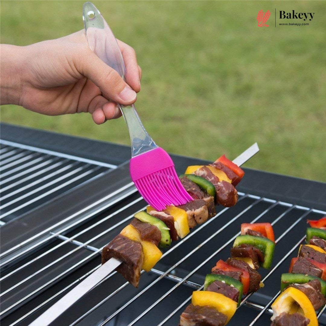 A hand uses a pink silicone basting brush with a clear handle to brush oil onto grilled skewers of meat and vegetables on a black grill outdoors, with a green grassy background and a Bakeyy logo in the top right corner.