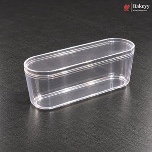 A clear plastic oval-shaped container with a matching lid, shown on a dark textured surface. The Bakeyy logo is visible in the top right corner.