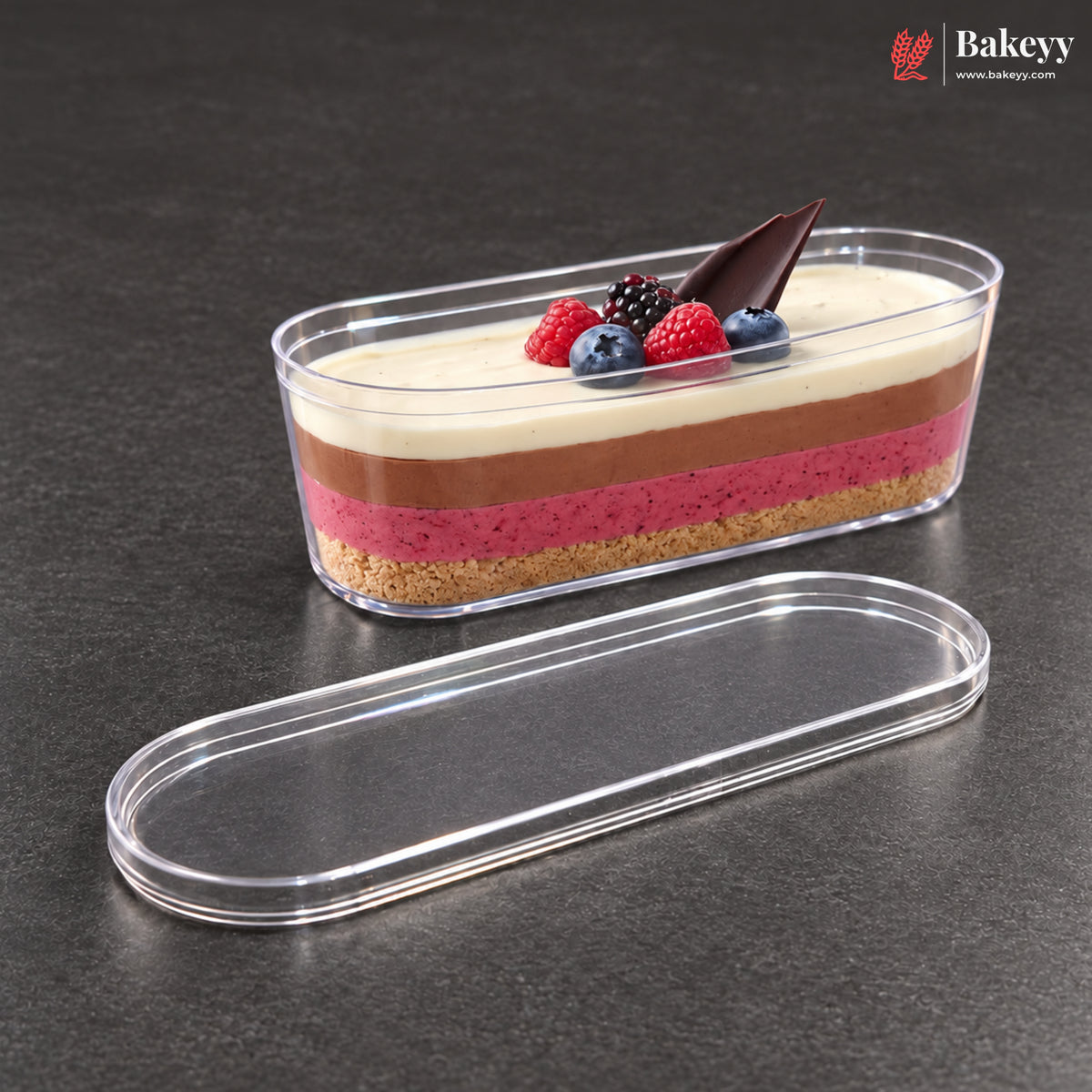 A clear plastic oval dessert container holding a layered cheesecake with cream, chocolate, raspberry, and graham cracker layers, topped with berries and a chocolate garnish, shown with its matching clear lid on a dark gray surface. The Bakeyy logo appears in the top right corner.