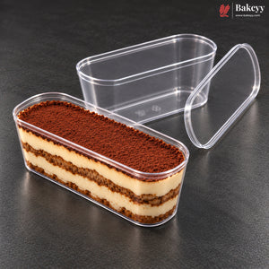 Clear plastic rectangular food container with a matching lid, shown on a dark gray surface. One container is open and filled with a layered dessert topped with cocoa powder, while the other container in the background is empty and transparent.