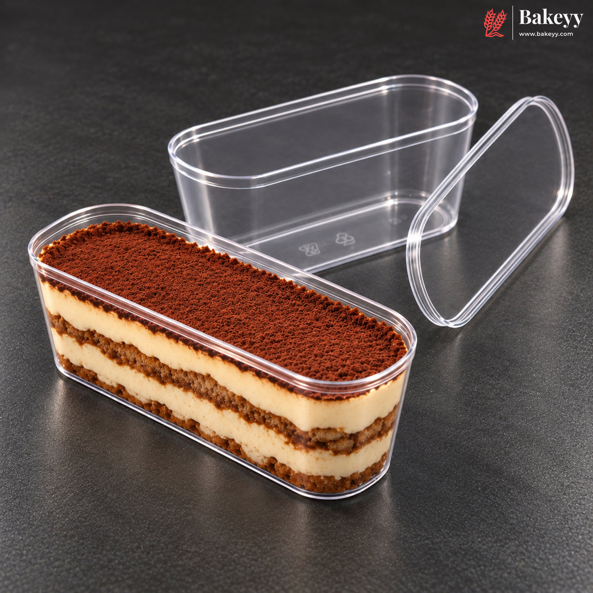 Clear plastic rectangular food container with a matching lid, shown on a dark gray surface. One container is open and filled with a layered dessert topped with cocoa powder, while the other container in the background is empty and transparent.