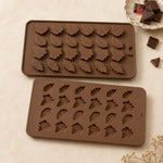Two brown silicone molds with leaf-shaped cavities, shown on a light beige surface with chocolate pieces and dried flowers as props.