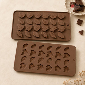 Two brown silicone molds with leaf-shaped cavities, shown on a light beige surface with chocolate pieces and dried flowers as props.
