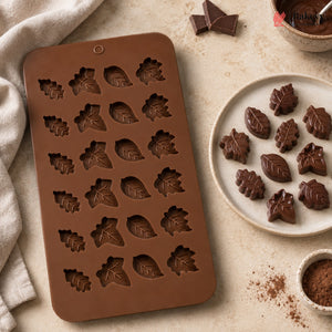 A brown silicone mold with leaf-shaped cavities, shown on a beige surface beside chocolate pieces and a plate of molded chocolate truffles.
