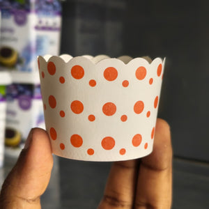 A hand holding a white scalloped paper cupcake liner decorated with orange polka dots, with blurred product packaging in the background.