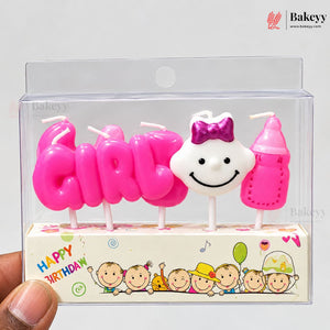 A boxed birthday candle set shaped like the word 'girl' in bright pink, a white smiling face with a purple bow, and a pink baby bottle candle, shown in a clear plastic package with a printed base. A hand is holding the package at the lower left.