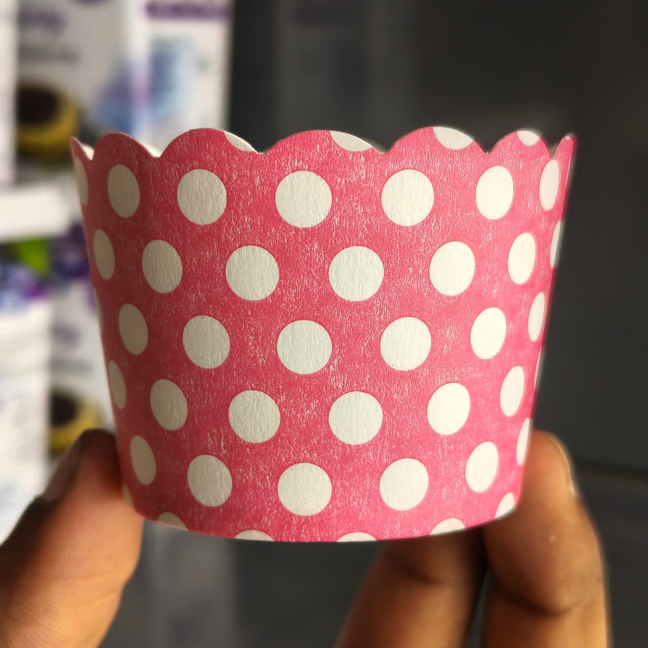 A pink paper cupcake liner with white polka dots and a scalloped edge, held by two hands against a blurred background.