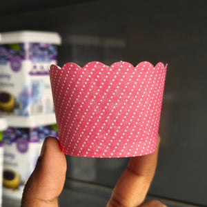 A pink scalloped paper cupcake liner with white polka dots, held by a person's fingers in front of a blurred background of packaged baked goods.
