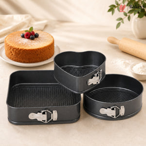 Three-piece black non-stick springform cake pan set with silver clamps, displayed on a beige fabric surface with baking props like a rolling pin, flour, and a decorated cake in the background.