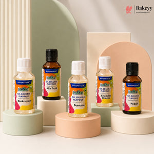Four bottles of Bakeyville oil soluble flavours arranged on pastel display pedestals against a clean studio backdrop. The bottles include Makkurrim, Banana, Cheddar Cheese, and Peach flavours, with two amber glass bottles and two clear plastic bottles, each labeled with the Bakeyville logo.