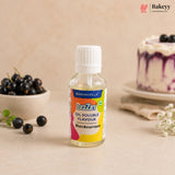 A bottle of Bakersville Uzley Blackcurrant Oil Soluble Flavour, set on a beige surface with fresh blackcurrants and a decorated cake in the background.