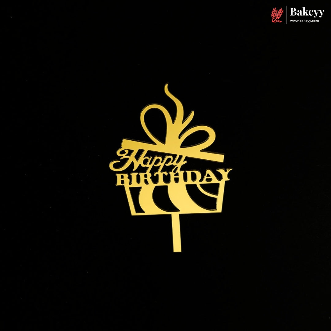 A gold cake topper shaped like a gift box with a bow and the words 'Happy Birthday' on a black background. A small Bakeey logo appears in the top right corner.