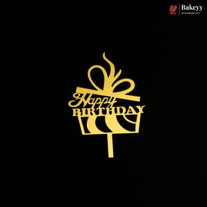 A gold cake topper shaped like a gift box with a bow and the words 'Happy Birthday' on a black background. A small Bakeey logo appears in the top right corner.