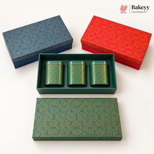 Three decorative rectangular boxes with matching lids are arranged on a white background. One box is open and contains three small cylindrical green tins with gold patterns.