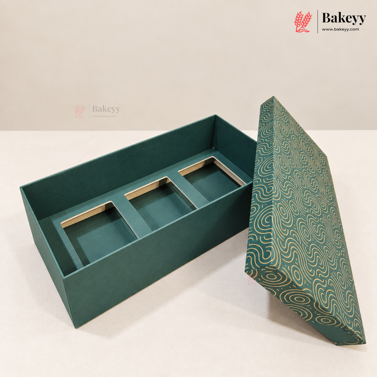 A rectangular teal gift box with three interior compartments and a patterned lid leaning against it on a light neutral surface. The lid has a swirling wave design in teal and cream.