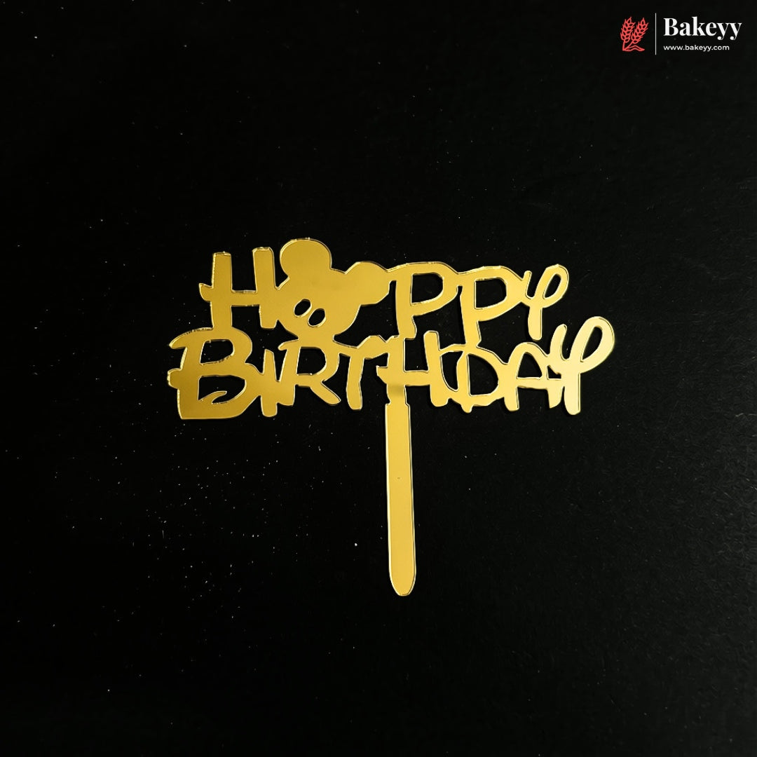 A gold-colored 'HAPPY BIRTHDAY' cake topper with a Mickey Mouse head silhouette, displayed on a black background.