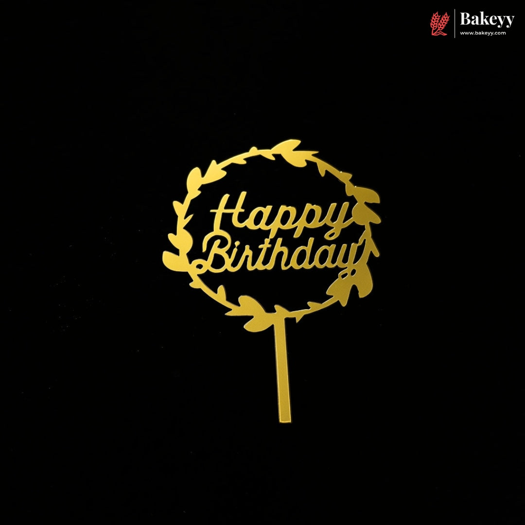 A gold metal 'Happy Birthday' cake topper with a leafy wreath design on a stick, shown against a solid black background. A Bakeey brand logo is visible in the top right corner.
