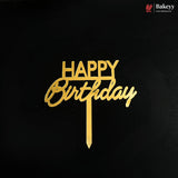 Gold metal 'HAPPY Birthday' cake topper with a vertical stick, displayed on a solid black background. A small 'Bakeyy' brand logo is visible in the top right corner.