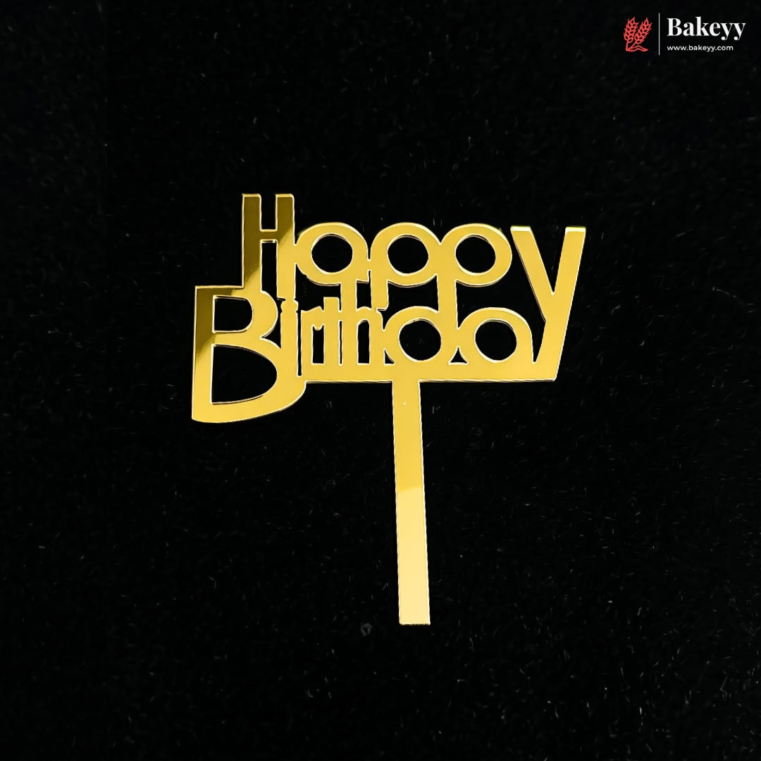 A gold-colored metal 'Happy Birthday' cake topper with a straight stem, shown on a textured black background. A 'Bakeey' brand logo is visible in the top right corner.