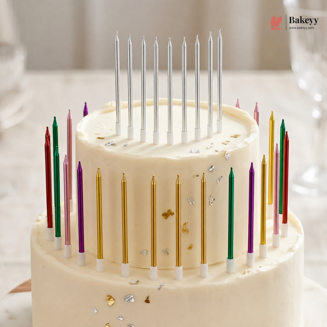 A two-tier cream cake decorated with colorful birthday candles and small metallic confetti, photographed on a softly blurred table setting. A Bakeyy logo appears in the top right corner.