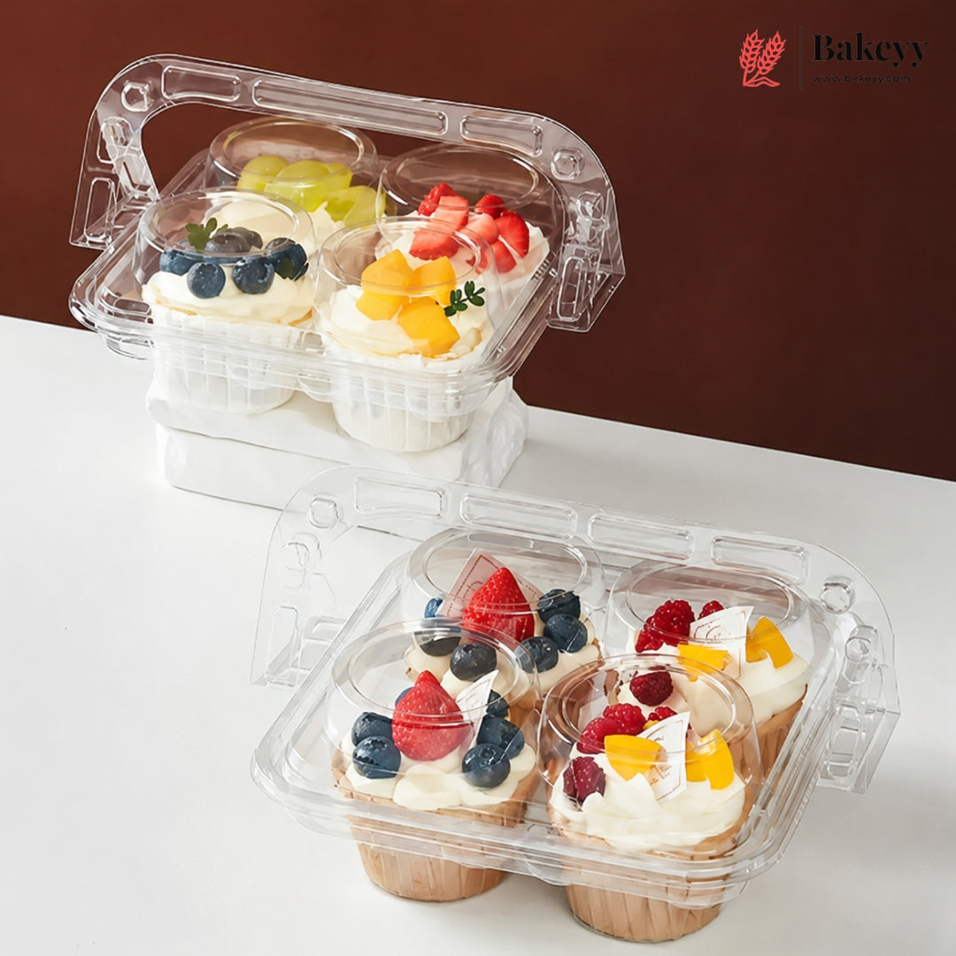 Two clear plastic cupcake containers with transparent lids, shown on a white surface against a dark brown background. The top container holds four frosted cupcakes with blueberries, green grapes, strawberries, and mango pieces.