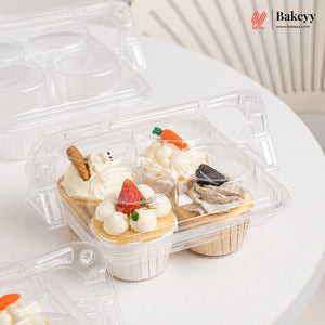 Clear plastic cupcake packaging tray holding four decorated cupcakes on a white round table, with a Bakeyy logo watermark in the top right.