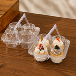 Two clear plastic cupcake carrier trays with triangular handles, one empty and one holding two decorated cupcakes, set on a wooden tabletop.