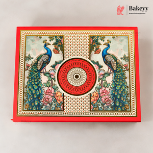 A decorative rectangular baking box with a red border and an ornate printed lid featuring two peacocks, floral motifs, and a central circular medallion. The Bakeyy logo appears in the top right corner.