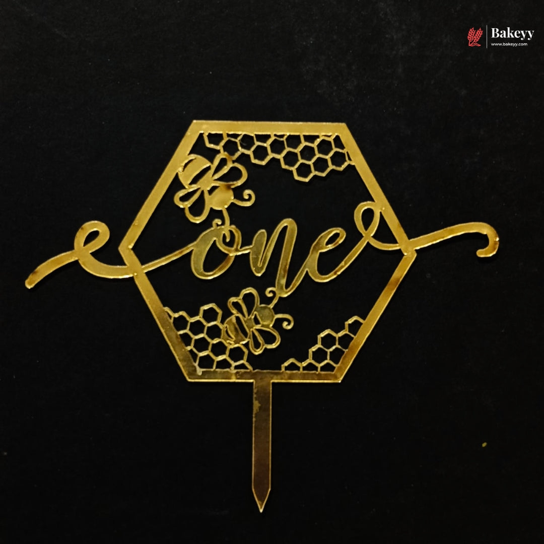 A gold metallic hexagonal cake topper with honeycomb patterns, bees, and the word 'one' in script, shown on a black background. A Bakeey watermark is visible in the top right corner.