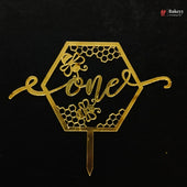 A gold metallic hexagonal cake topper with honeycomb patterns, bees, and the word 'one' in script, shown on a black background. A Bakeey watermark is visible in the top right corner.