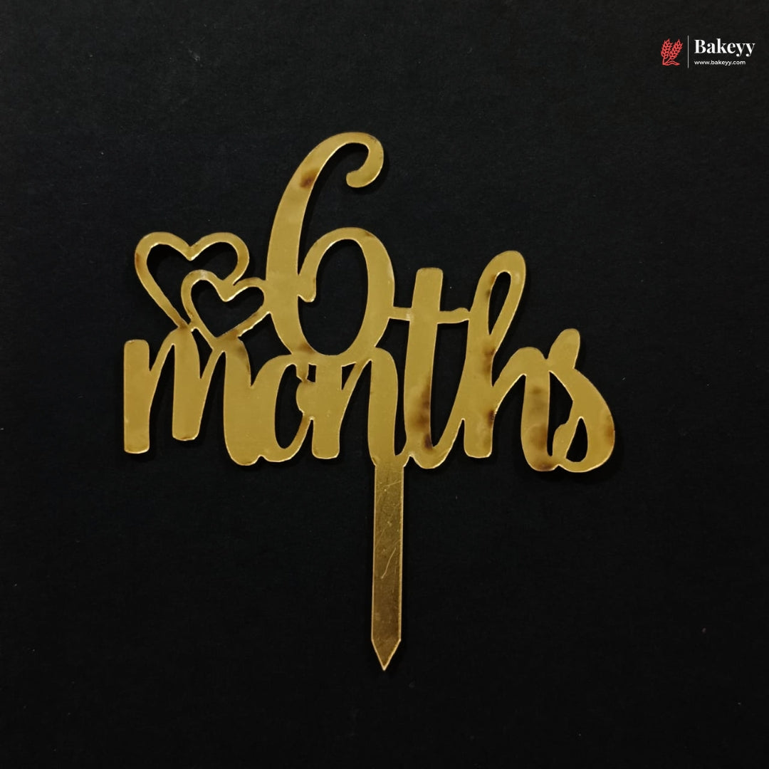 A gold metallic 6 months cake topper with two heart shapes on a black background, featuring a Bakeyy brand watermark in the top right corner.