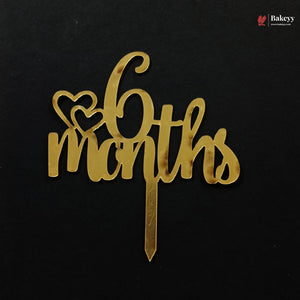 A gold metallic 6 months cake topper with two heart shapes on a black background, featuring a Bakeyy brand watermark in the top right corner.