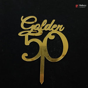 A gold metallic 'Golden 50' cake topper with a single wire stake, shown on a solid black background. A small 'Bakeyy' brand watermark is visible in the top right corner.