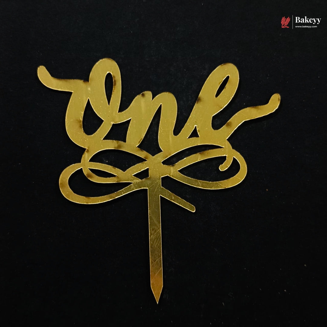 A gold metallic script cake topper spelling 'one' with a decorative bow motif and a straight pick, shown on a black background.