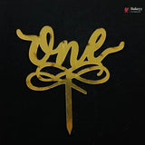 A gold metallic script cake topper spelling 'one' with a decorative bow motif and a straight pick, shown on a black background.