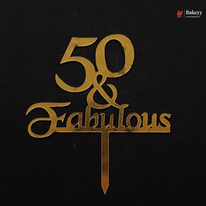 Gold metal cake topper with the text '50 & Fabulous' on a black background, featuring a small Bakeey watermark in the top right corner.