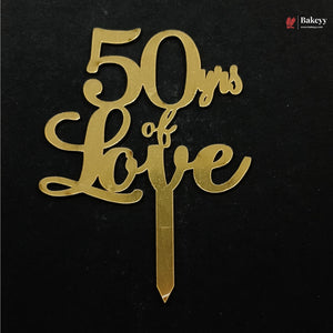 Gold metallic cake topper with the text '50 yrs of Love' in elegant cursive script, complete with a stake, shown on a black background.