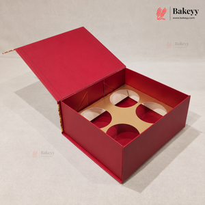 A red bakery gift box with its lid open, showing a beige cardboard insert with circular cutouts. The box is photographed on a neutral light gray background with Bakeyy branding visible.
