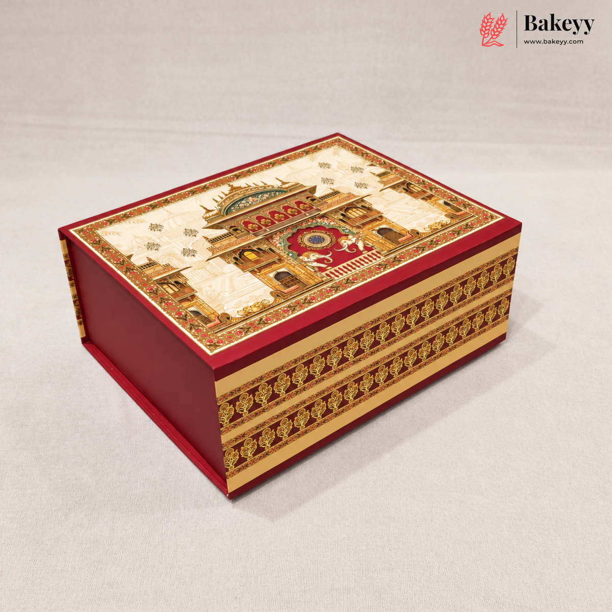 A decorative red and gold gift box with an intricate architectural illustration on the lid, photographed on a light gray background. The Bakeyy logo is visible in the top right corner.
