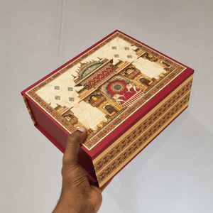 A hand holding a rectangular decorative gift box with a red border and an illustrated central scene in beige, gold, and red tones, photographed against a plain light background.