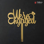 Gold metal 'We are Engaged' cake topper with a ring icon, shown on a black background with a Bakeey brand watermark in the top right corner.