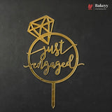 A gold metal 'just engaged' cake topper with a diamond graphic and stake, displayed on a dark textured background. A Bakeyy logo is visible in the top right corner.